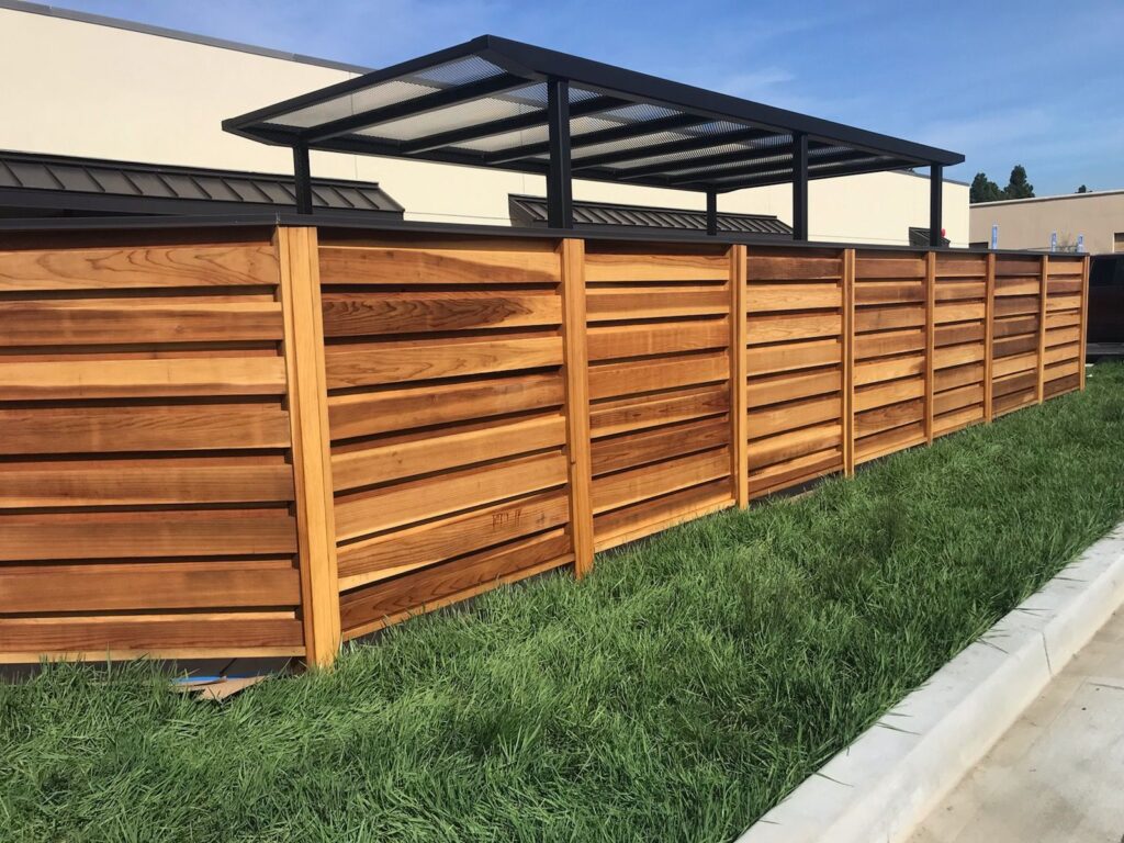 A wooden horizontal slat fence with a covered structure behind it, bordered by grass and a concrete curb.