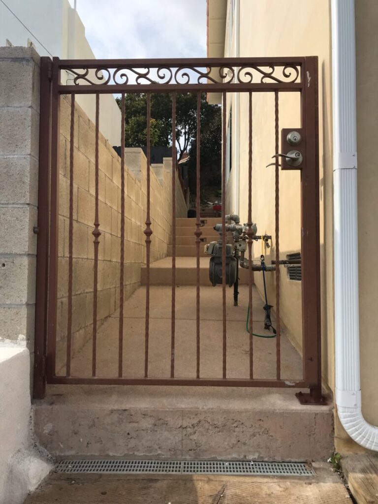 A brown metal gate with decorative top opens to a narrow pathway between concrete walls, leading to stairs and utility meters.